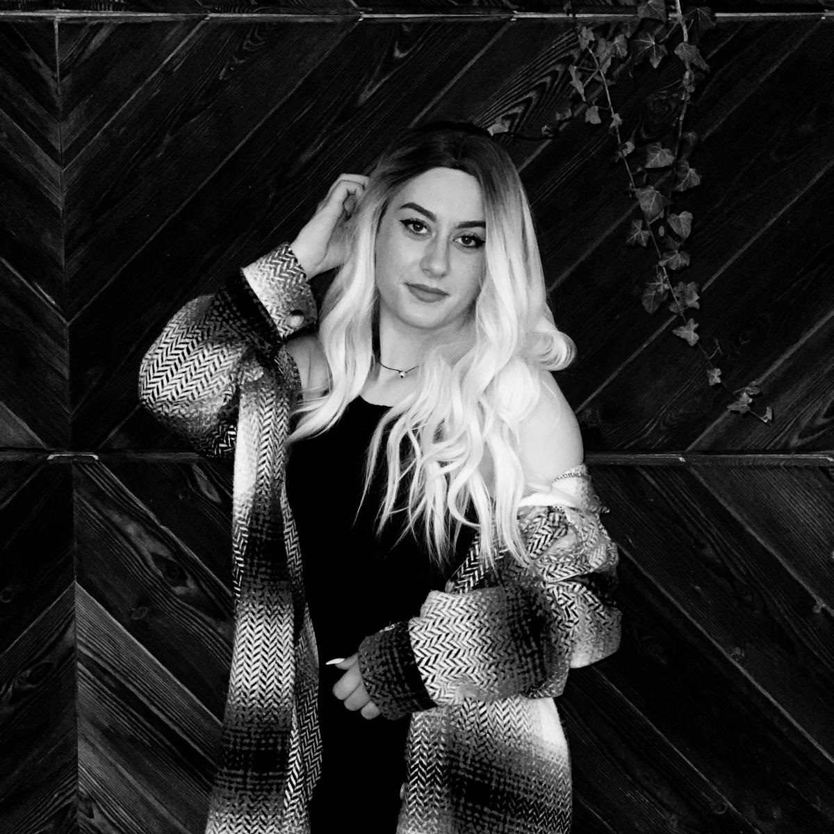 Porträt einer Frau mit langen, blonden Haaren in einem gestreiften Outfit vor einer Holzwand.